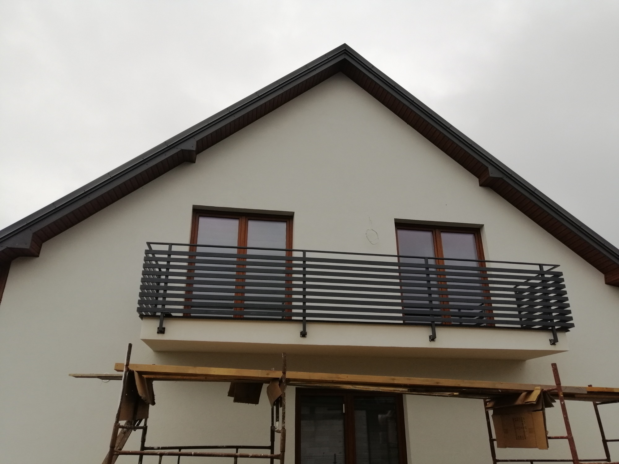 Nowoczesna balustrada balkonowa z poziomych, czarnych profili, zamontowana na białej elewacji domu z brązowymi ramami okiennymi, widoczna z dołu ze stojącym rusztowaniem.