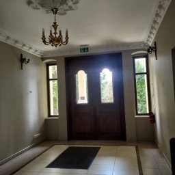 Gabinet Specjalistyczny Agnieszka Izbicka. Psycholog, Dietetyk, Psychodietetyk - Wnętrze holu z drewnianymi drzwiami i dekoracyjnym sufitem z żyrandolem. Widoczne płytki podłogowe, okna po bokach drzwi i gaśnica na ścianie. Klasyczny wystrój.