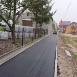 Nowo wyasfaltowana droga osiedlowa z krawężnikami, ogrodzeniem panelowym i stosem kostki brukowej, ubijana walcem drogowym w oddali.