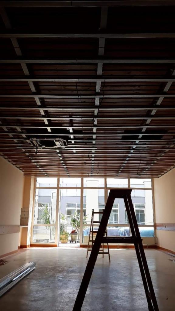 Montaż metalowej konstrukcji sufitu podwieszanego w jasnym pomieszczeniu z oknami, widoczna drabina i materiały budowlane na podłodze.