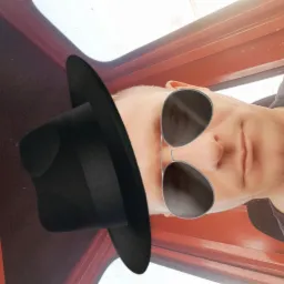 Autoportret mężczyzny w kapeluszu i okularach przeciwsłonecznych, z nietypowej perspektywy, w kabinie maszyny budowlanej. Czerwone elementy wnętrza kabiny.