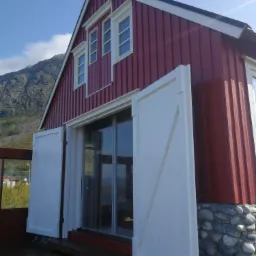 Czerwony, drewniany dom z białymi okiennicami na kamiennej podstawie, z widokiem na górę i błękitne niebo.