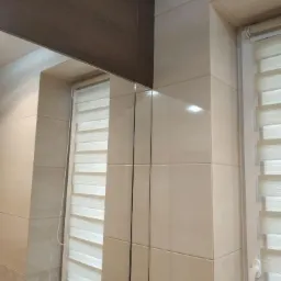 Wykończona łazienka z beżowymi, błyszczącymi płytkami ceramicznymi, brązowym pasem dekoracyjnym pod sufitem i roletami okiennymi typu dzień-noc.