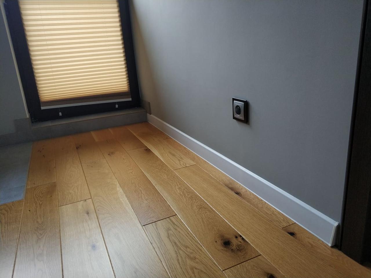 Parkiet z jasnego drewna ułożony w jodełkę, szara ściana z czarnym gniazdkiem elektrycznym i białą listwą przypodłogową, okno z żaluzjami plisowanymi.