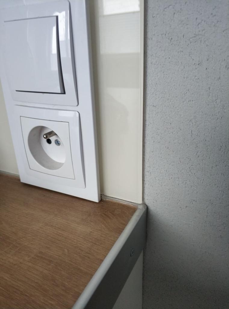 Biały włącznik światła i gniazdko elektryczne zamontowane na ścianie obok blatu kuchennego z okleiną drewnopodobną, przylegające do białej ściany z połyskiem i szarej ściany o fakturze baranka.