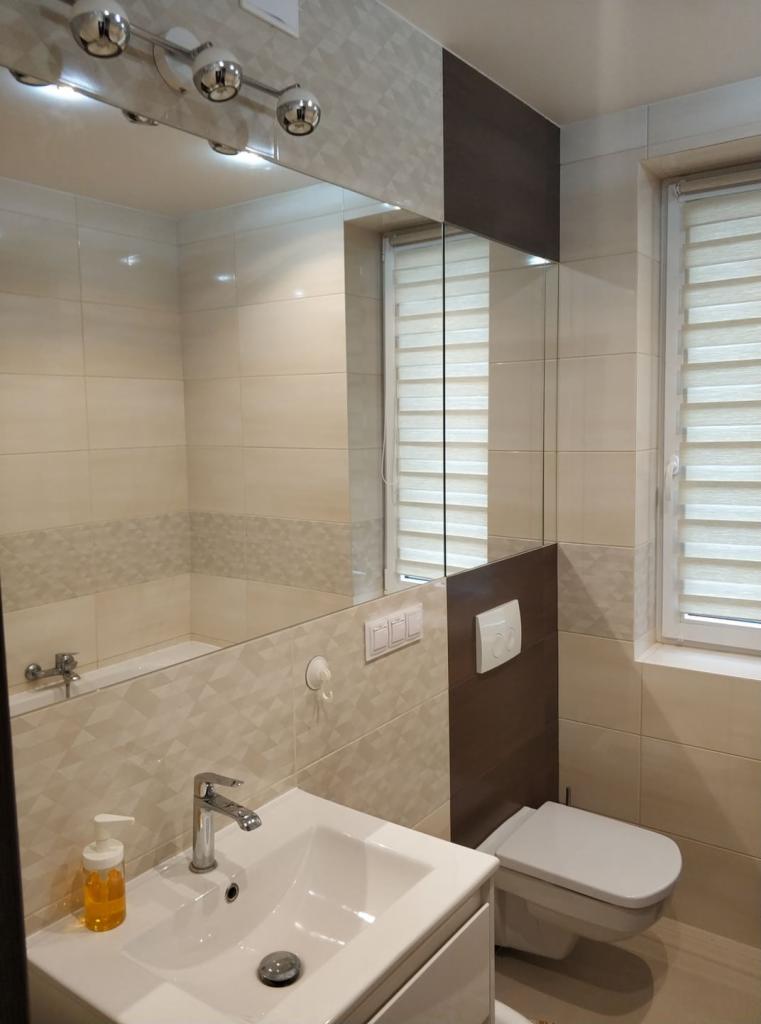 Beżowa łazienka z umywalką, toaletą i dużym lustrem, fragment wanny widoczny w odbiciu, dekoracyjne płytki na ścianach, rolety w oknie.