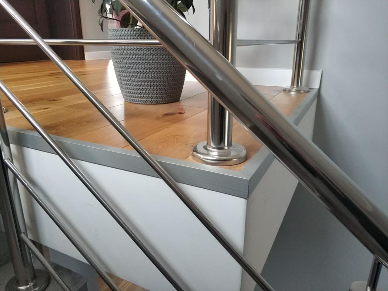 Szczegółowe ujęcie nowoczesnej, metalowej balustrady schodowej z drewnianymi stopniami, uzupełnionej o donicę z rośliną w tle.