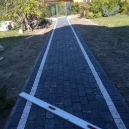 Świeżo ułożona ścieżka z ciemnej kostki brukowej z białymi obrzeżami prowadząca do furtki w ogrodzie we Włocławku, widoczna poziomica.