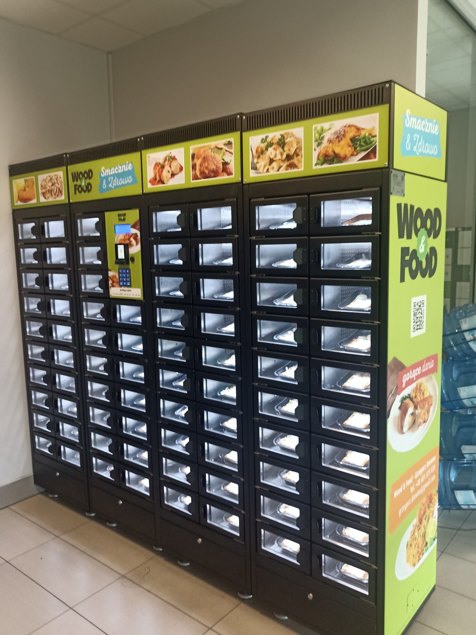 Automat vendingowy z gotowymi daniami firmy Wood Food, z widocznymi zdjęciami potraw i panelem dotykowym do wyboru posiłków.