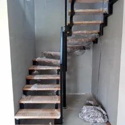 Wnętrze w trakcie wykańczania: schody z drewnianymi stopniami zabezpieczonymi folią i czarną metalową konstrukcją, widoczne przewody elektryczne i materiał zabezpieczający podłogę.