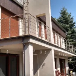 Nowoczesny balkon z prostą, nierdzewną balustradą z poziomych prętów, widok z dołu na jasny budynek z brązowymi okiennicami i wiszącymi ozdobnymi żarówkami.