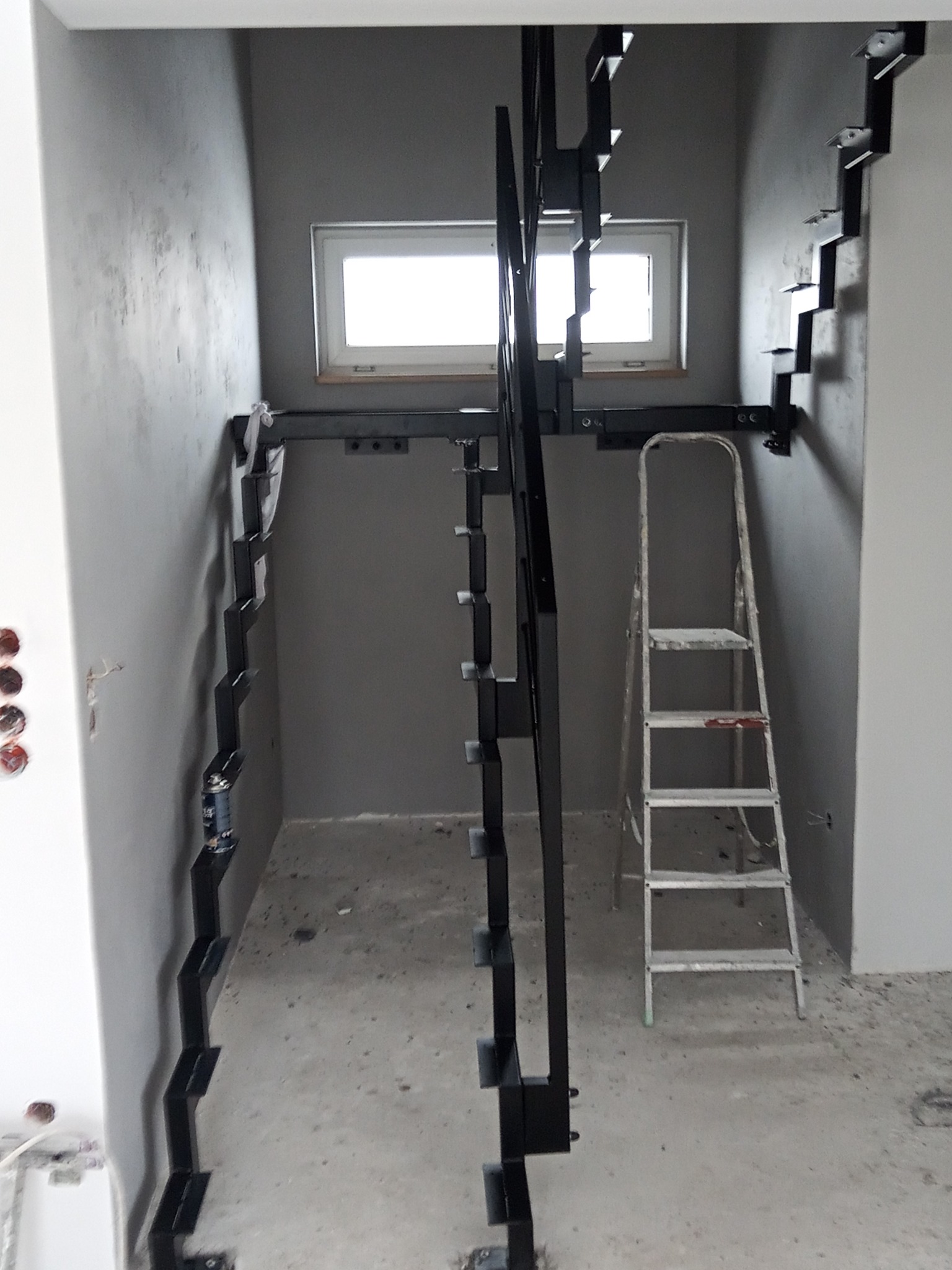 Montaż schodów metalowych w surowym wnętrzu, widoczne wsporniki schodów przymocowane do ściany i metalowa konstrukcja oraz drabina malarska.