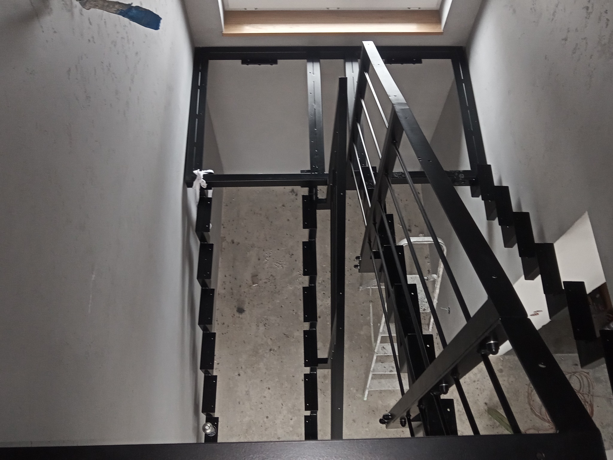 Wysoki kąt widzenia na częściowo zmontowaną, czarną konstrukcję schodów metalowych z minimalistyczną balustradą, widoczne ślady po malowaniu na ścianie i drabina w tle.
