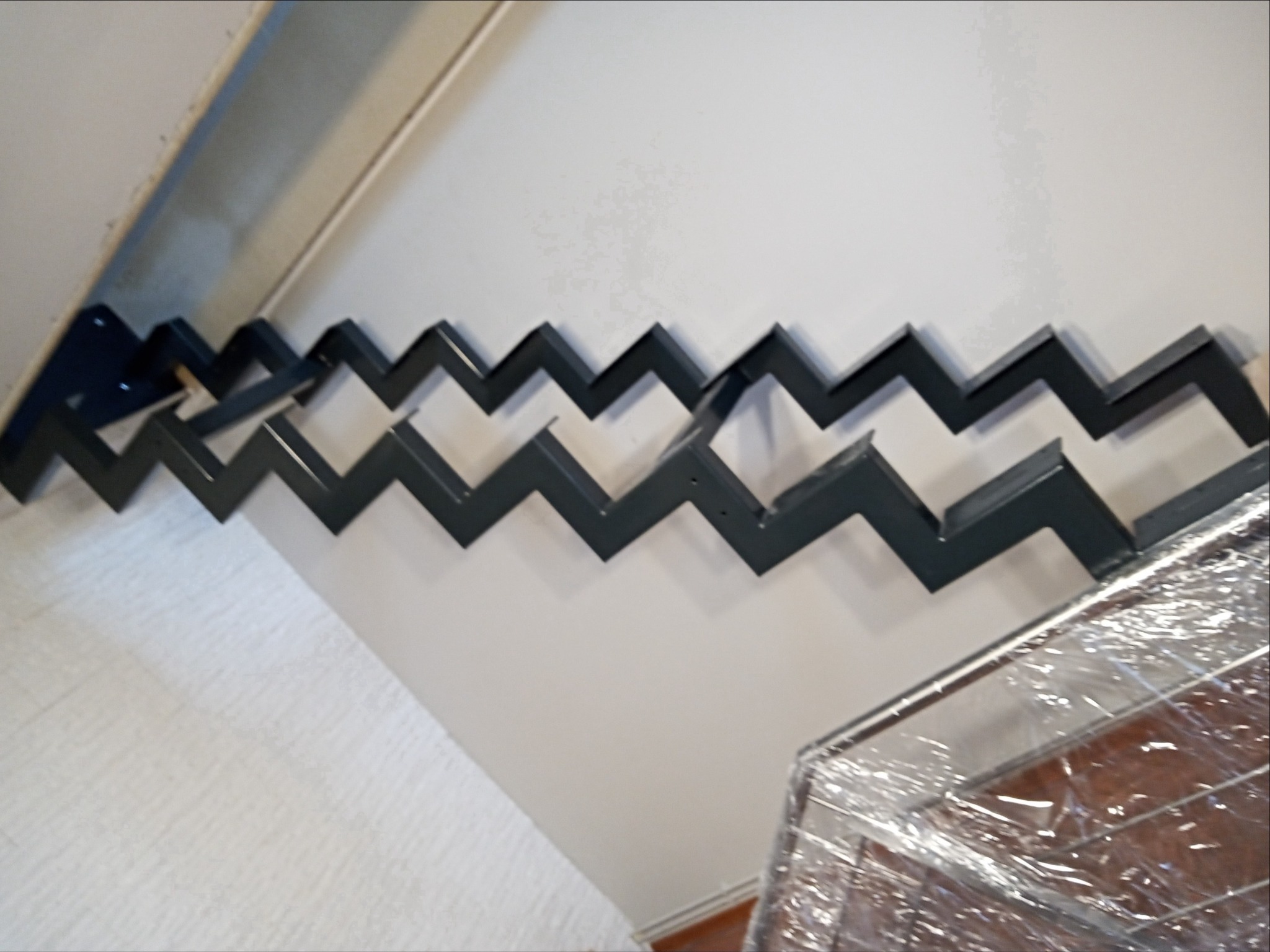 Zygzakowate, metalowe elementy konstrukcyjne przymocowane do ściany, prawdopodobnie fragment schodów lub balustrady w trakcie montażu, widoczne zabezpieczone folią elementy.