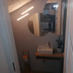 Niewielka łazienka z umywalką nablatową na drewnianym blacie, okrągłym lustrem i nietypowym oświetleniem sufitowym, widok zza otwartych drzwi.