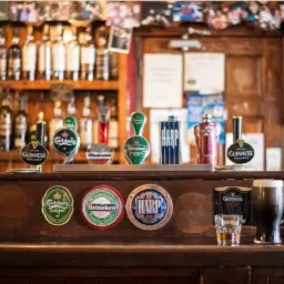 Bar z drewnianym blatem, nalewaki piwa różnych marek (Guinness, Harp, Heineken, Budweiser, Carlsberg) oraz szklanka ciemnego piwa i kieliszek alkoholu.