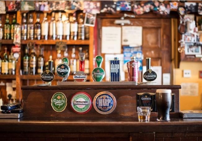 Bar z drewnianym blatem, nalewaki piwa różnych marek (Guinness, Harp, Heineken, Budweiser, Carlsberg) oraz szklanka ciemnego piwa i kieliszek alkoholu.