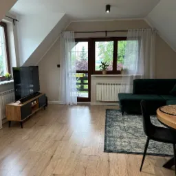 Jasne wnętrze poddasza z drewnianą podłogą, telewizorem na szafce RTV, zieloną sofą narożną i widokiem na ogród przez przeszklone drzwi balkonowe z białymi zasłonami.