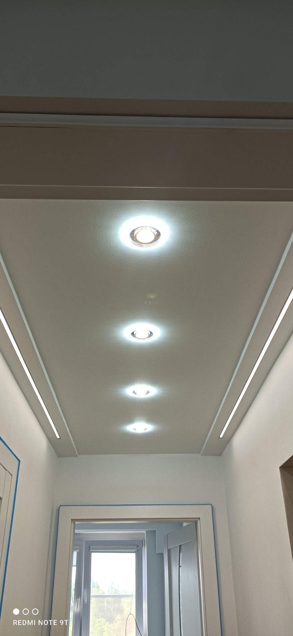 Biały, podwieszany sufit z wbudowanymi okrągłymi lampami punktowymi i liniowym oświetleniem LED po bokach w nowocześnie wykończonym wnętrzu.