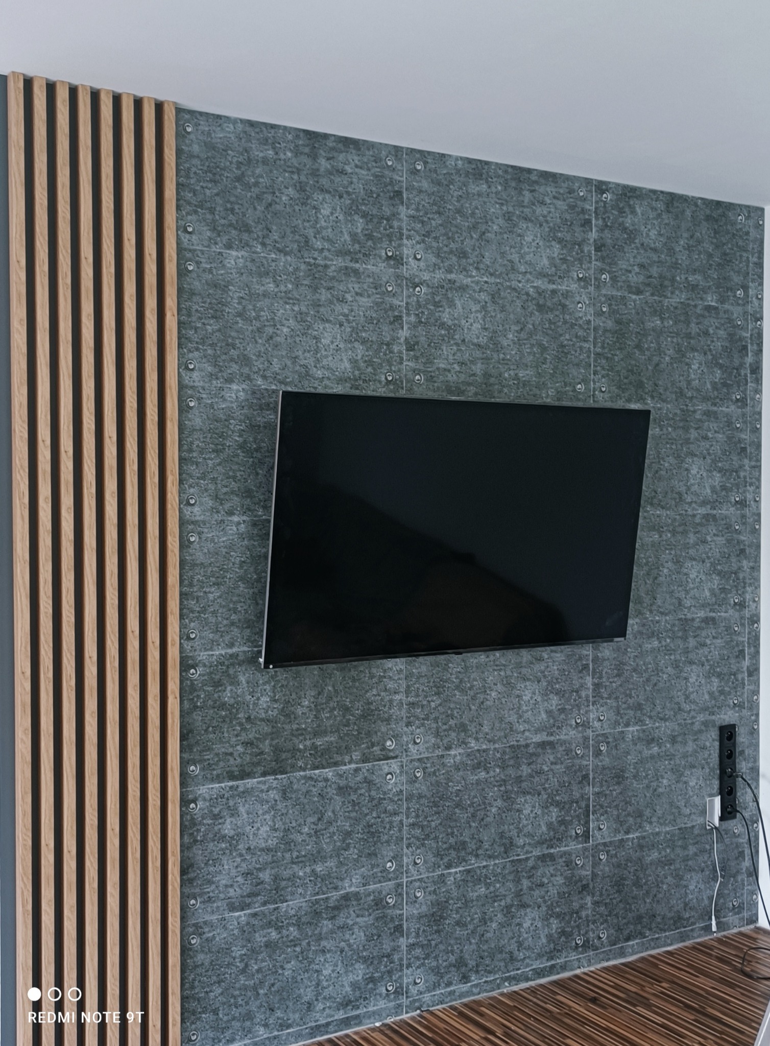 Szara ściana z płytek imitujących metal, ozdobiona drewnianymi lamelami i telewizorem; widoczne gniazdka elektryczne z wtyczkami.