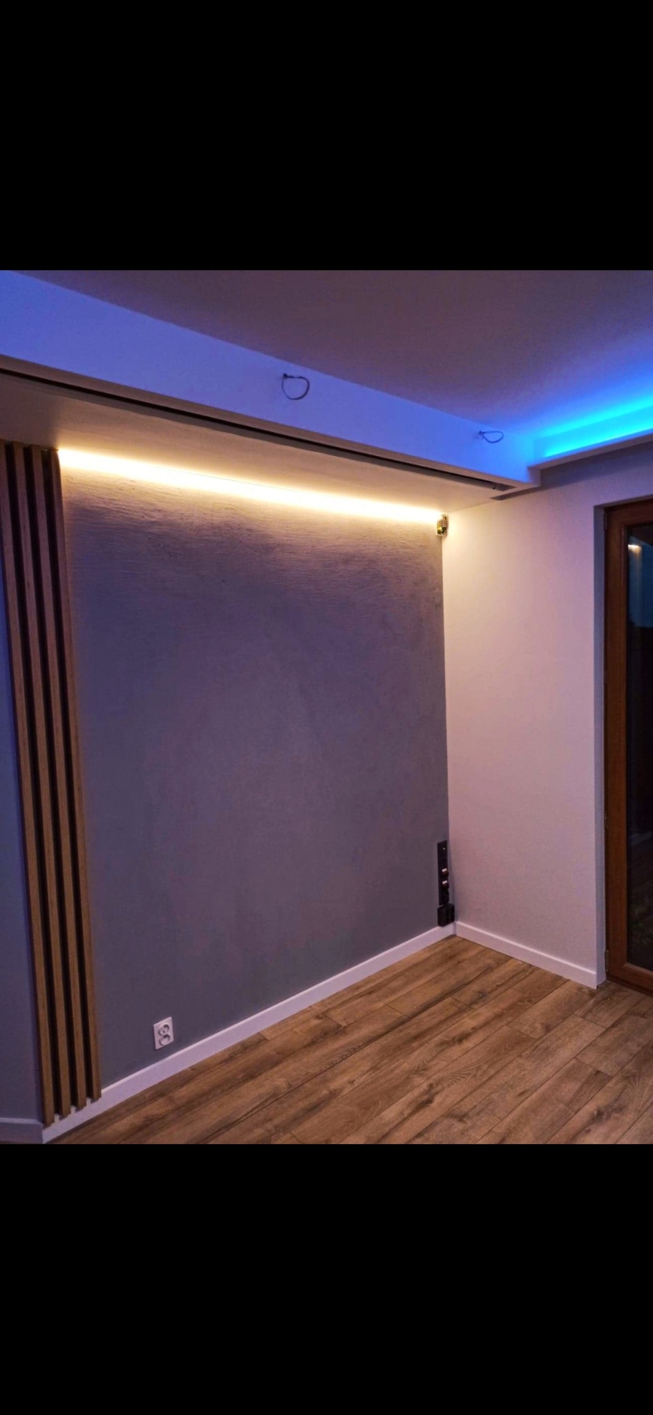 Szara ściana z dekoracyjnymi drewnianymi panelami i podświetleniem LED w Piastowie, drewniana podłoga, niebieskie oświetlenie sufitowe.