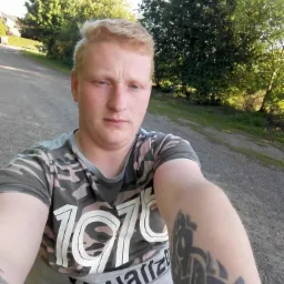 Młody mężczyzna z tatuażem na ramieniu pozuje na tle zieleni i drogi. Ubrany w koszulkę moro z napisem. Widoczne drzewa i polna droga.