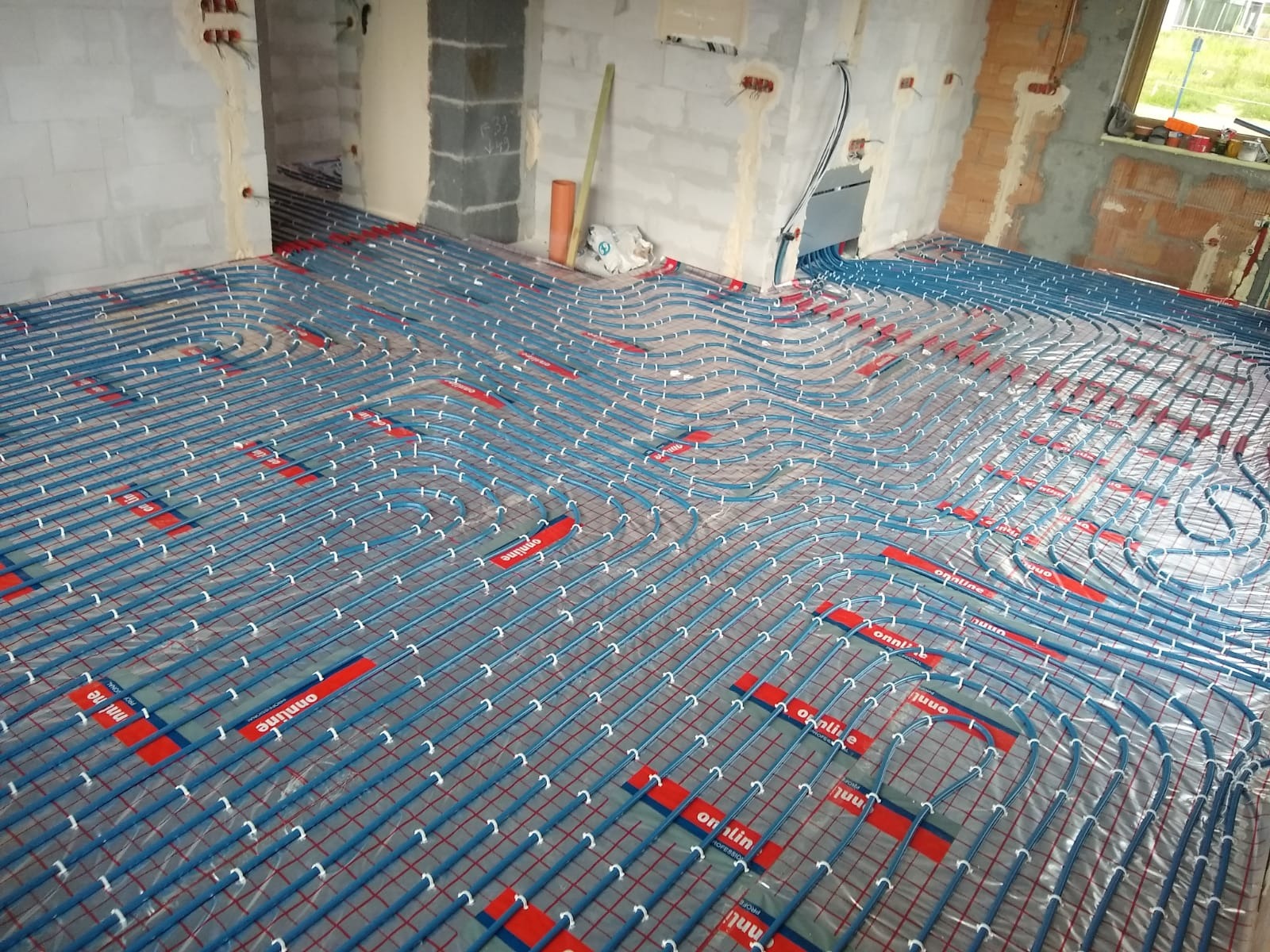 Rozłożona instalacja ogrzewania podłogowego w surowym stanie, rurki w kolorze niebieskim na siatce z tworzywa, widoczne fragmenty ścian i przygotowane otwory instalacyjne.