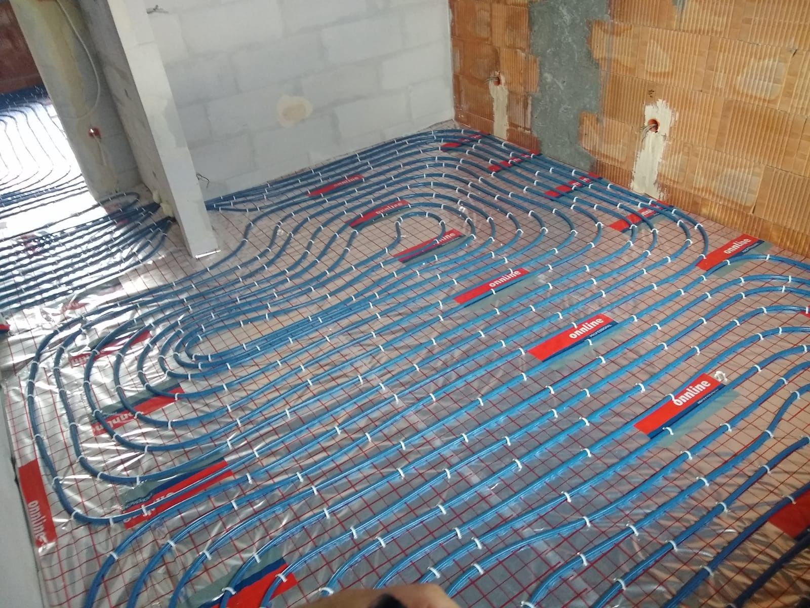 Niebieskie rury ogrzewania podłogowego ułożone na siatce w surowym wnętrzu, widoczne fragmenty ścian z cegły i betonu.