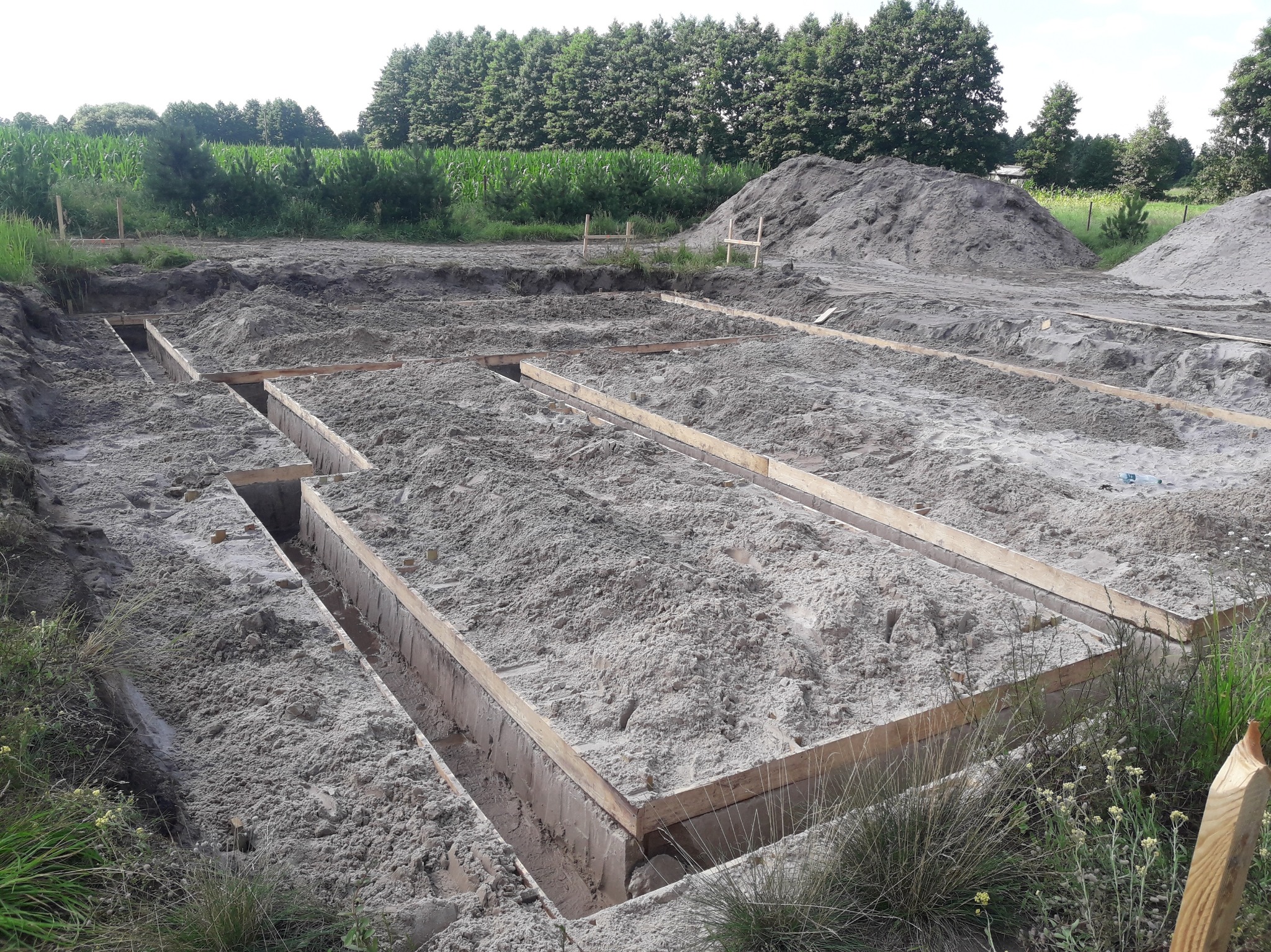 Przygotowanie fundamentów pod dom murowany: wykopane rowy zalane piaskiem, drewniane szalunki w ziemi, hałdy piasku w tle, widok z góry na plac budowy.