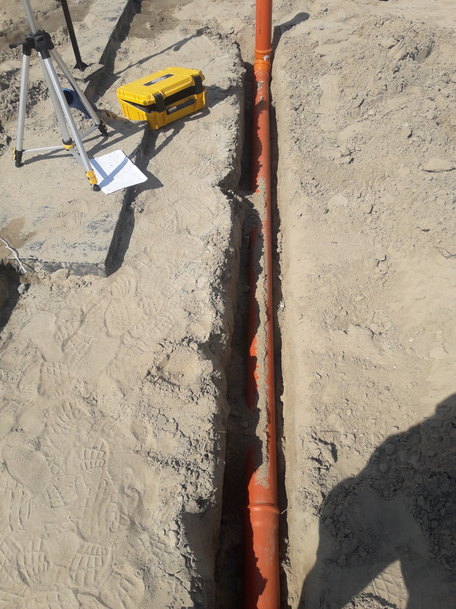 Pomarańczowe rury ułożone w wykopie w ziemi, obok widoczny niwelator na statywie i żółta skrzynka narzędziowa, liczne ślady butów w piasku.