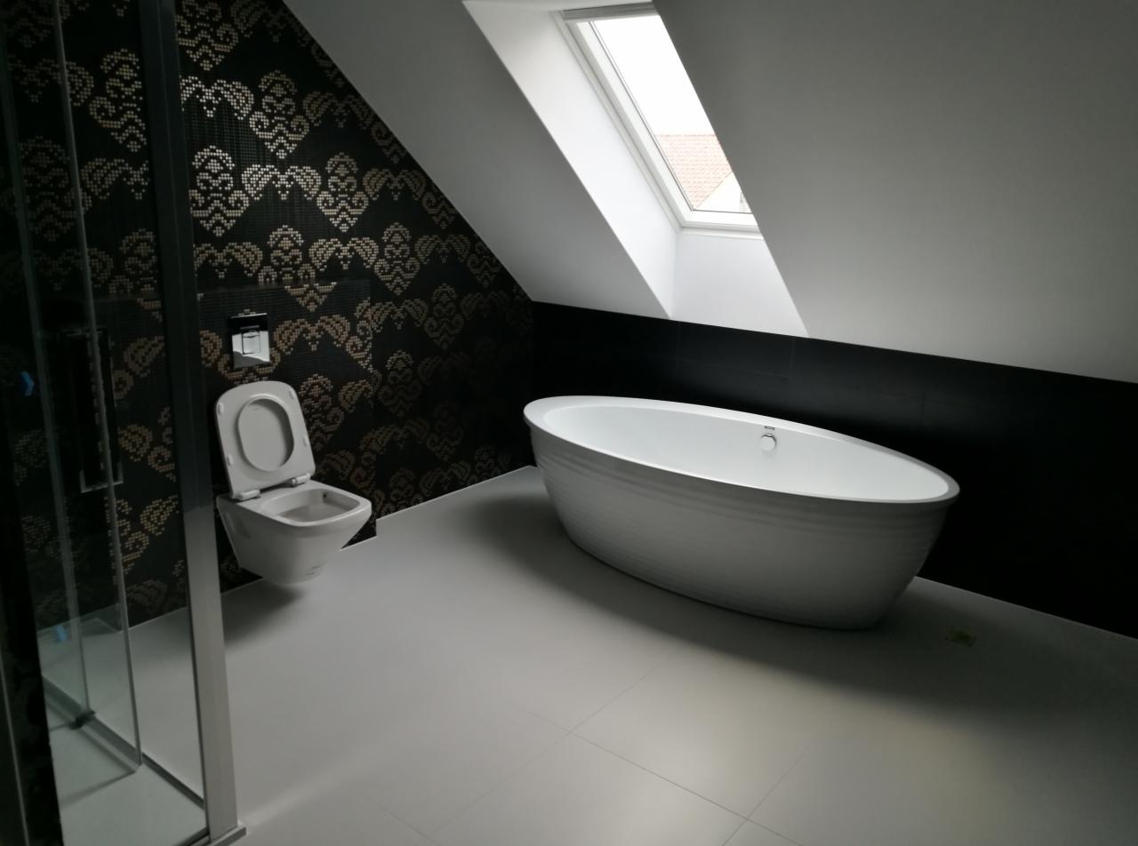 Nowoczesna łazienka na poddaszu z białą wanną wolnostojącą, podwieszaną toaletą i czarno-złotą mozaiką na ścianie, oświetlona światłem z okna dachowego.