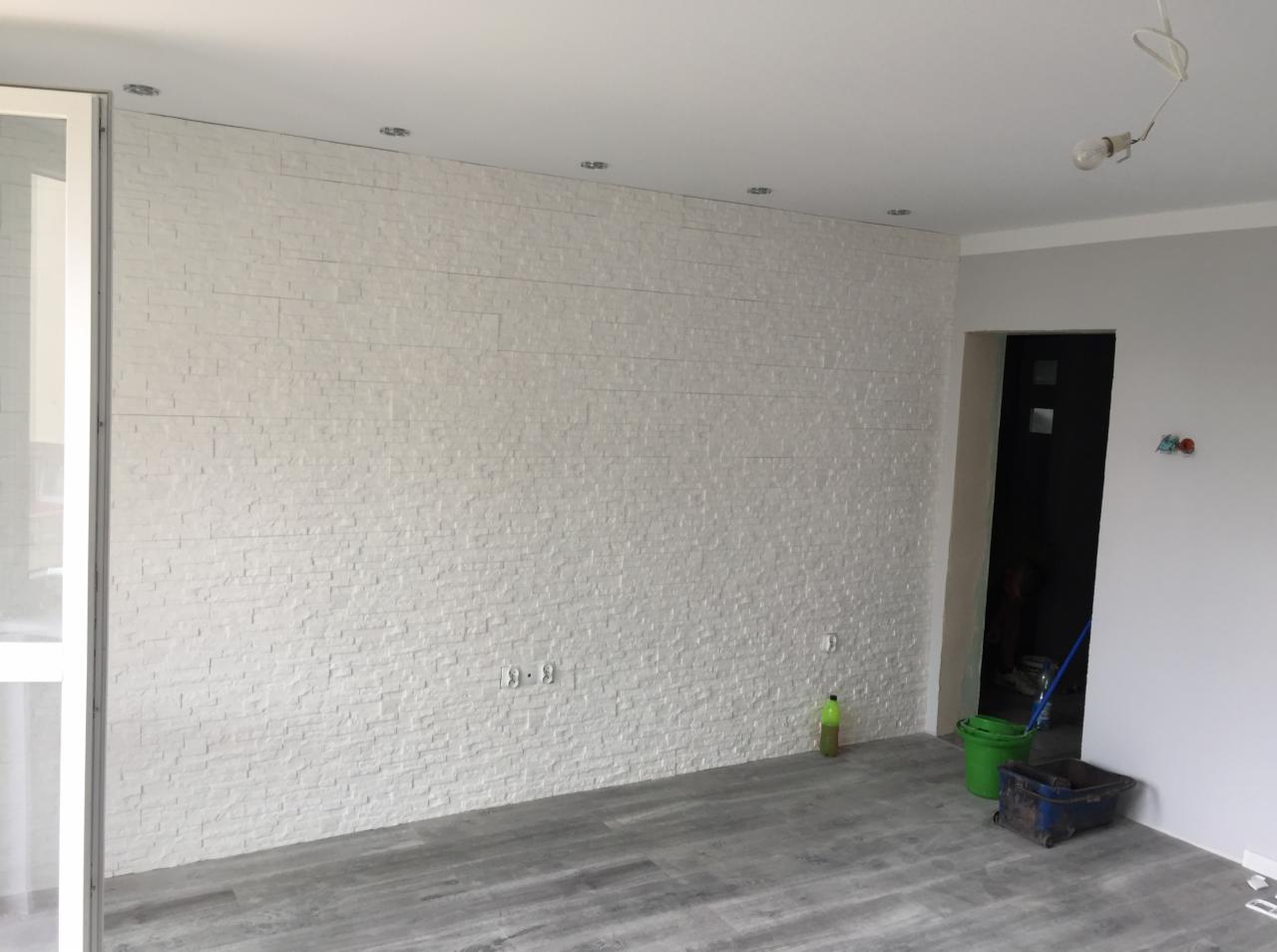 Ściana wykończona dekoracyjną białą okładziną imitującą kamień, z widocznymi gniazdkami elektrycznymi, szarą podłogą, wiadrem i mopem w tle, sugerującymi prace wykończeniowe.