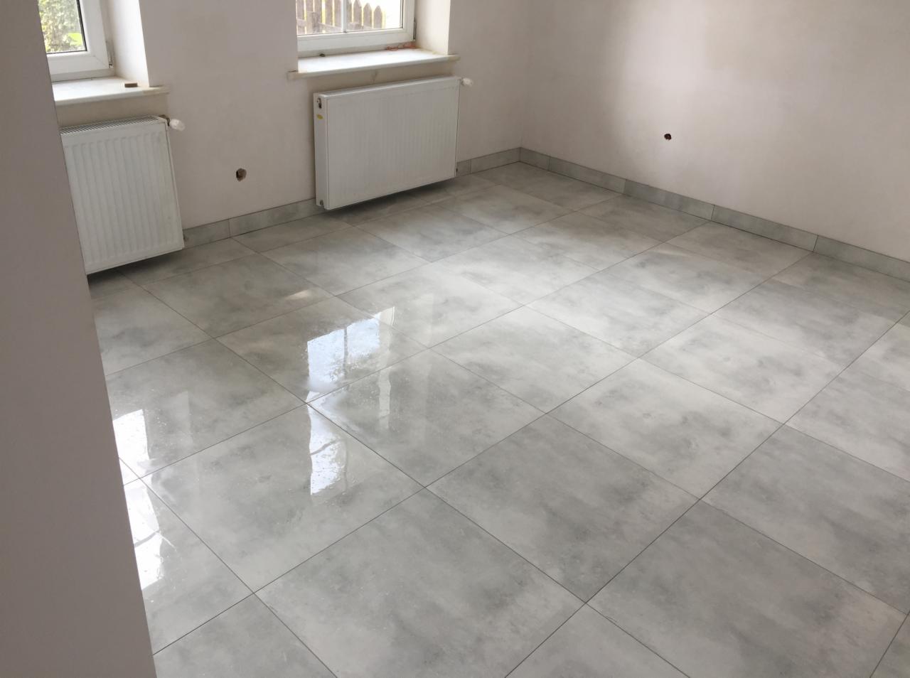 Wykończona podłoga z szarych, kwadratowych płytek w pomieszczeniu z dwoma grzejnikami i oknami. Widoczne odbicie światła w polerowanej powierzchni płytek.