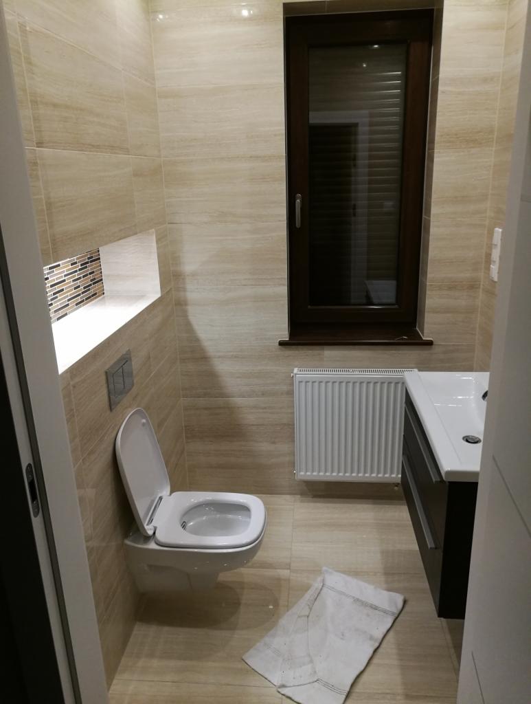 Ukończona łazienka z beżowymi płytkami imitującymi drewno, wisząca toaleta z otwartą klapą, grzejnik pod oknem z brązową ramą i biała umywalka z szafką w kolorze ciemnego brązu, widoczny ręcznik...