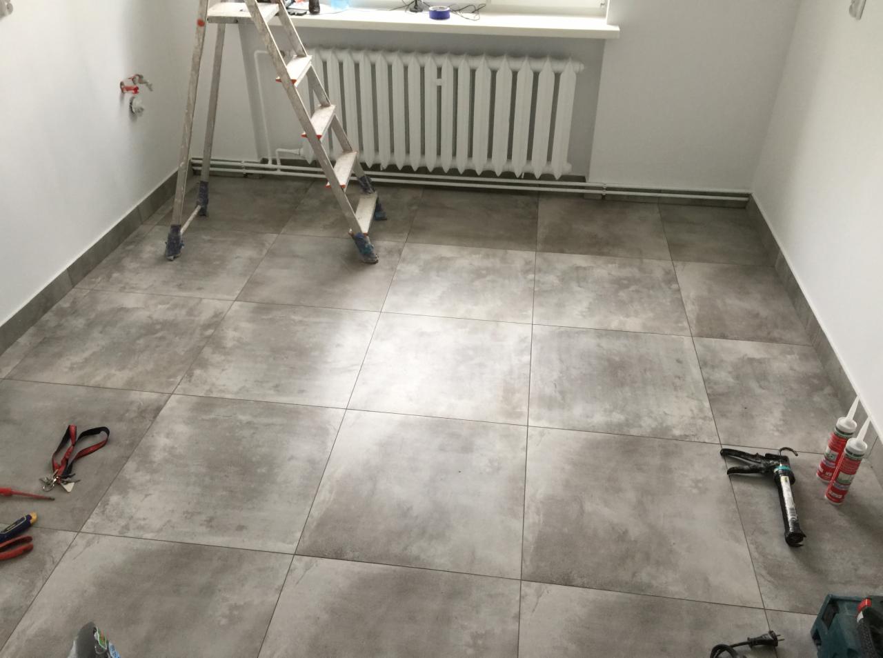 Szare, kwadratowe płytki podłogowe ułożone w pomieszczeniu z białymi ścianami, widoczna drabina, grzejnik, narzędzia glazurnicze i pistolet do silikonu.