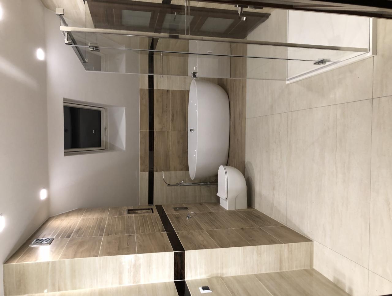 Nowoczesna łazienka z wolnostojącą wanną, kabiną prysznicową ze szklaną ścianką i toaletą w zabudowie; beżowe płytki imitujące drewno z czarnymi akcentami; małe okno w górnej części ściany.