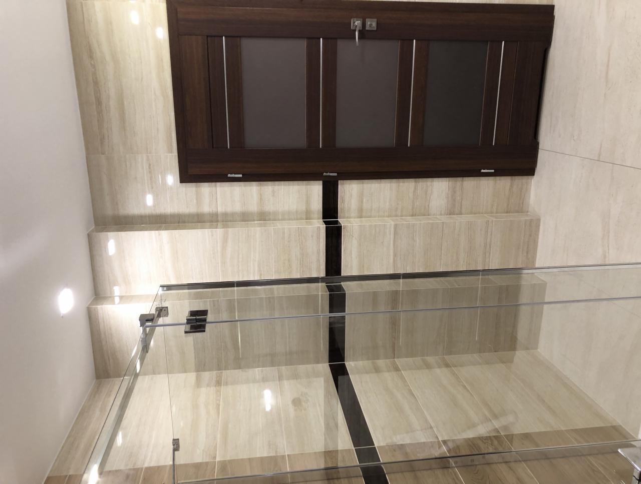 Wykończona łazienka z beżowymi płytkami imitującymi marmur, kabina prysznicowa ze szklanymi ścianami i ciemnobrązowa szafka wisząca z uchwytami.