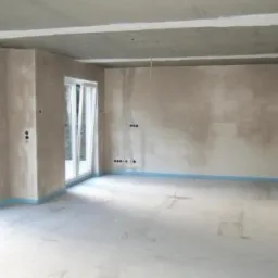 Wnętrze w stanie surowym: betonowa podłoga i ściany, okna przesuwne z widokiem na zieleń, widoczne instalacje elektryczne, zabezpieczenie folią wzdłuż ścian.