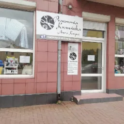 Fasada pracowni krawieckiej z witrynami wystawowymi prezentującymi uszyte kołdry, poduszki, zabawki i dekoracje, nad wejściem szyld z nazwą 'Pracownia Krawiecka Aneta Kropisz' oraz informacjami...