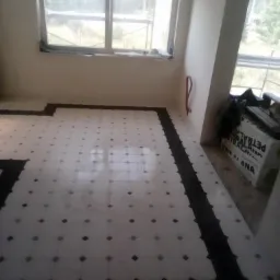 Układanie białych płytek podłogowych z czarnymi akcentami w pomieszczeniu z dwoma oknami, widoczne niezakończone krawędzie i kartony w tle.