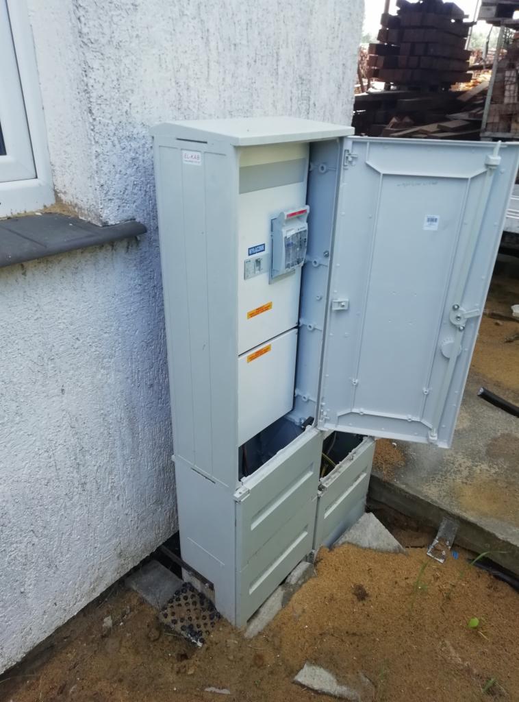 Otwarta skrzynka przyłączeniowa z licznikiem elektrycznym, zamontowana przy ścianie budynku, widoczne kable i elementy instalacji.