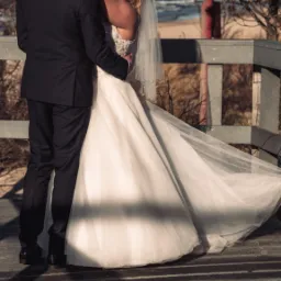 Uśmiechnięta panna młoda w koronkowej sukni i welonie obejmuje pana młodego na drewnianym pomoście z widokiem na morze i plażę.