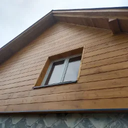 Szczyt domu z drewnianą elewacją w odcieniach brązu, z oknem na poddaszu, wykończony kamienną podmurówką w kształcie heksagonów.