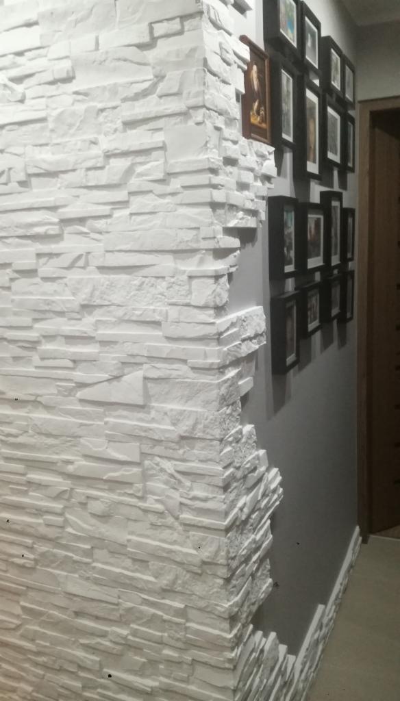Dekoracyjna ściana z białych kamieni imitujących cegłę, narożnik z niedokończonym wykończeniem, na szarej ścianie galeria zdjęć w czarnych ramkach.