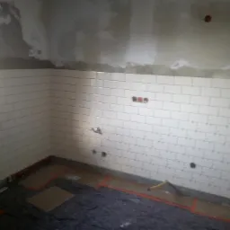 Fragment ściany w trakcie remontu kuchni, z ułożonymi białymi, prostokątnymi płytkami ceramicznymi na dolnej części ściany i surowym tynkiem powyżej; widoczne przygotowane punkty instalacji wodnej...
