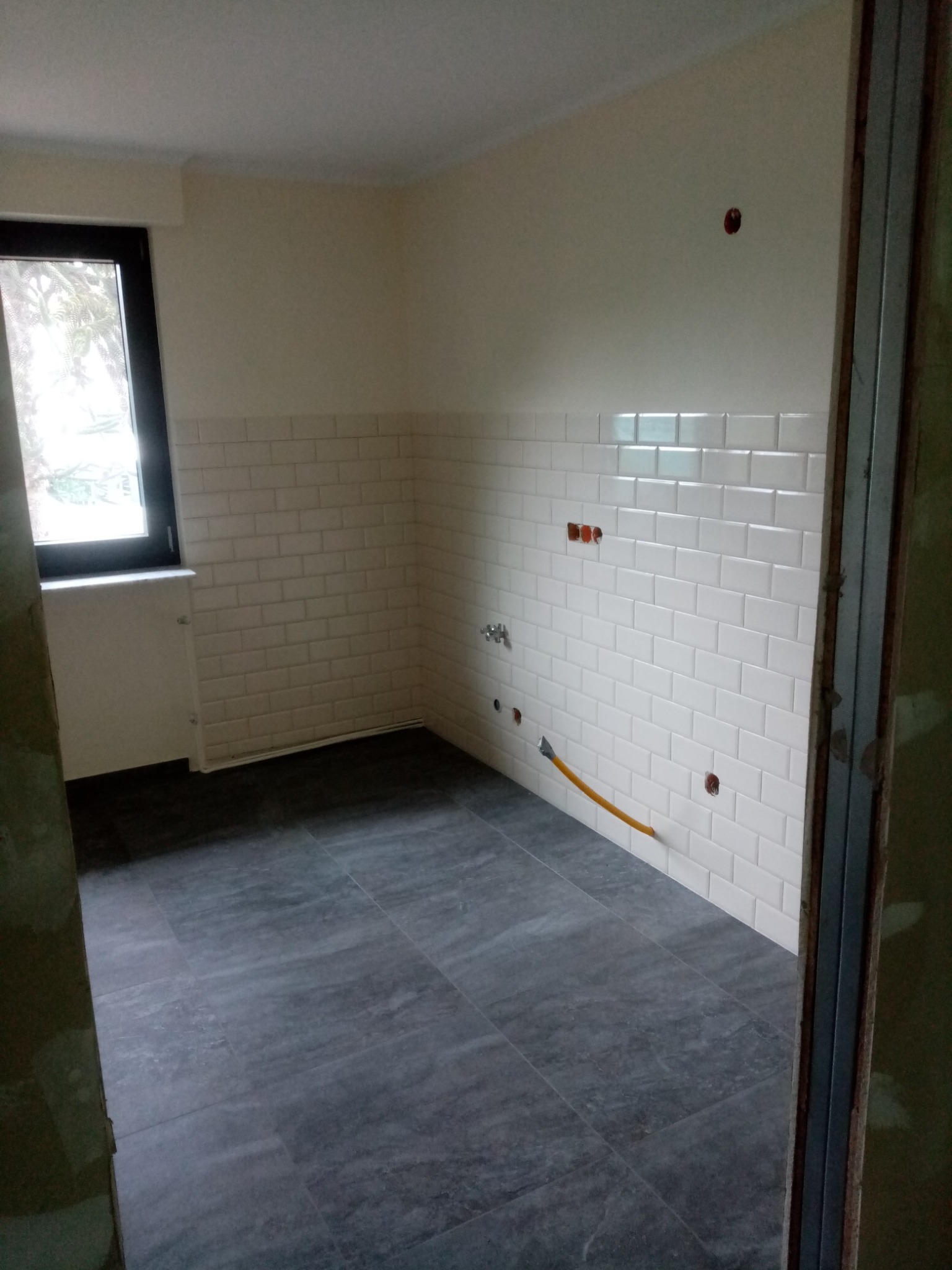 Wnętrze kuchni w trakcie remontu: ściany wyłożone białymi, prostokątnymi płytkami typu metro, szara podłoga z płytek, widoczne otwory na instalacje i żółty przewód, okno w czarnej ramie.