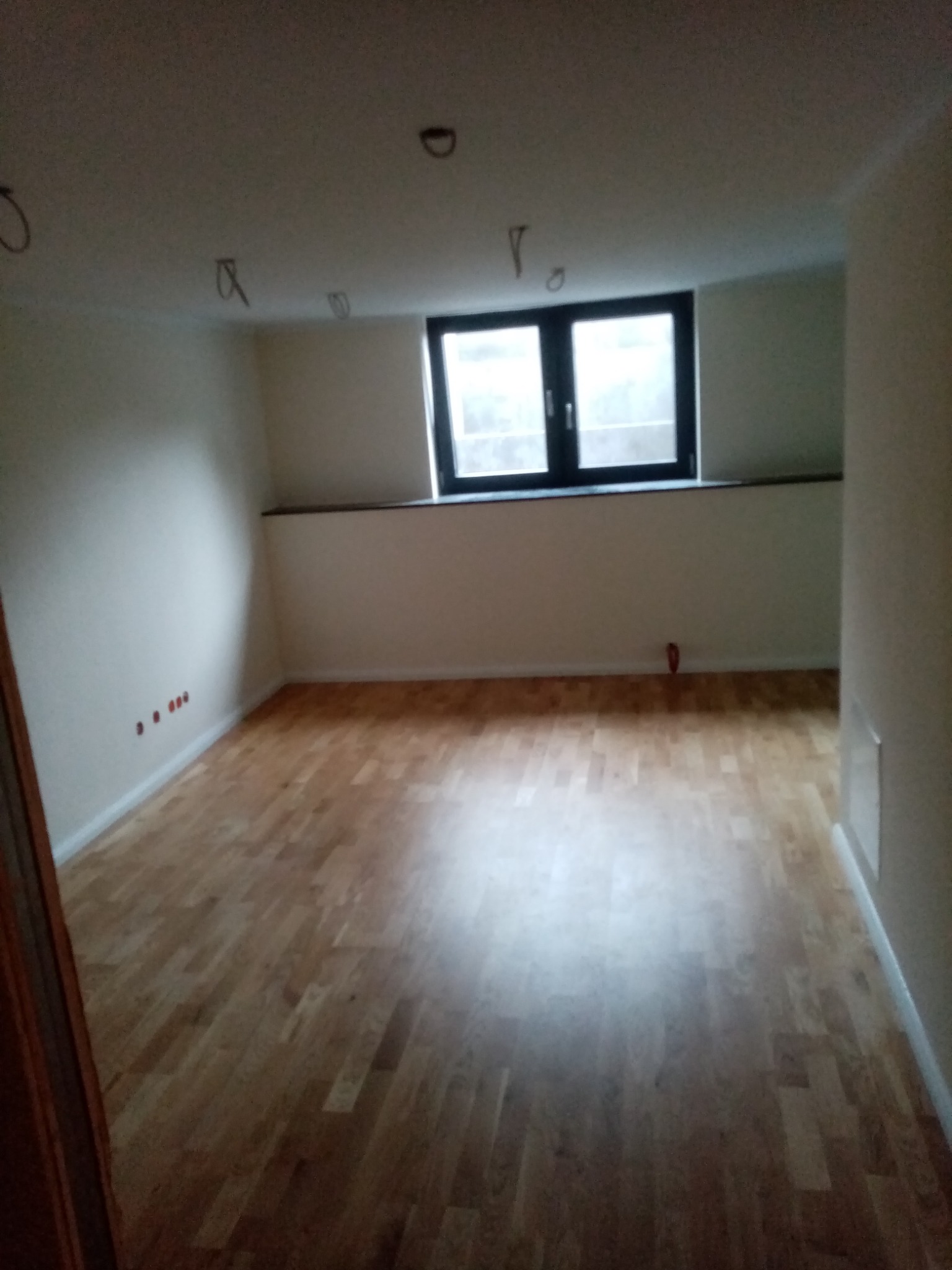 Pusty pokój z drewnianą podłogą, białymi ścianami i oknem. Widoczne wystające kable elektryczne ze sufitu oraz czerwone zaślepki gniazdek na ścianie.