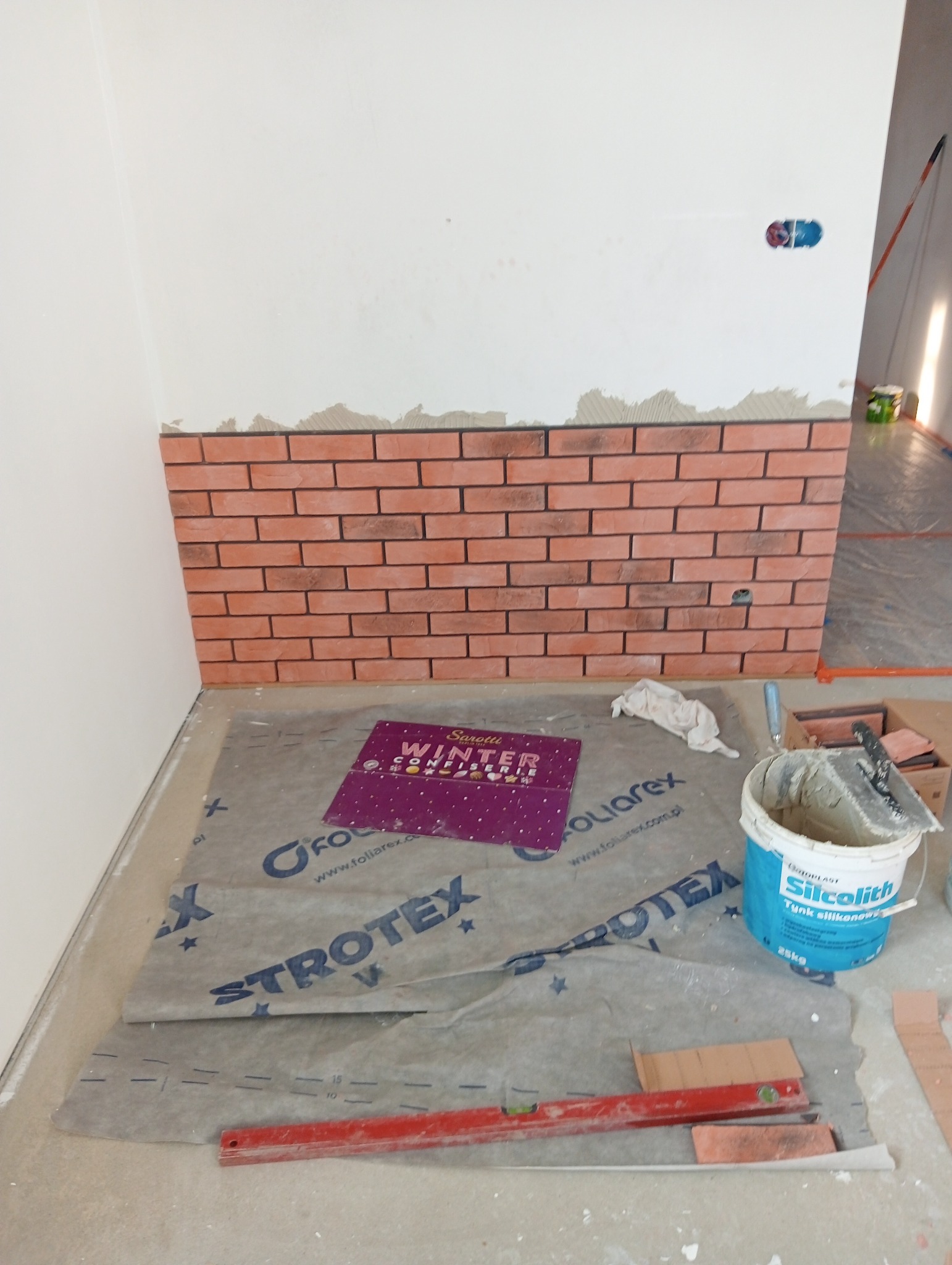 Fragment ściany z częściowo ułożoną dekoracyjną cegłą, wiadro z zaprawą Silcolith, poziomica i folia ochronna Strotex na podłodze.