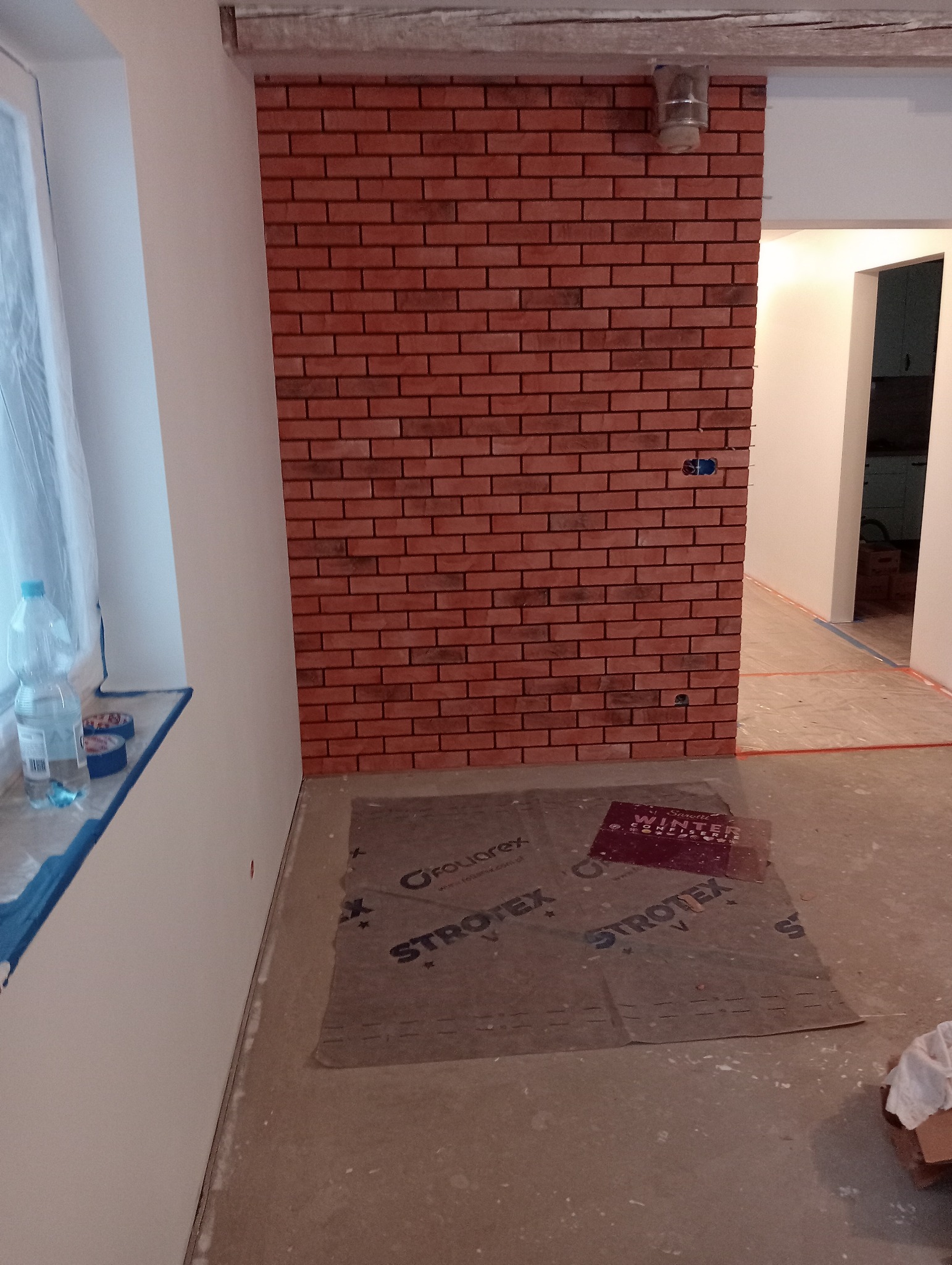 Wykończona ściana z cegły klinkierowej wewnątrz pomieszczenia, zabezpieczona podłoga folią ochronną Strotex, widoczne puszki elektryczne w ścianie i przygotowane pomieszczenie do dalszych prac...