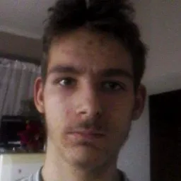 Portret młodego mężczyzny z krótkimi, ciemnymi włosami, patrzącego prosto w obiektyw, na tle jasnego wnętrza z widoczną szafką i firanką.